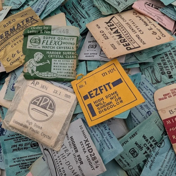 Collection of vintage watch crystal packaging labels on a textured surface.
